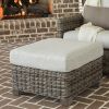 Woven wicker ottoman with light cushion in front of a chair and fireplace.