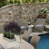 Laurent wicker dining side and arm chairs with cushions arranged around a stone outdoor dining table by a pool.