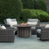 Trevi 50 inch round fire pit patio table in hickory finish surrounded by woven lounge chairs in landscaped outdoor patio